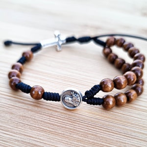 May include: A brown wooden bead bracelet with a silver cross charm and a silver medallion featuring a religious image. The bracelet is made with black string and has a double row of beads.