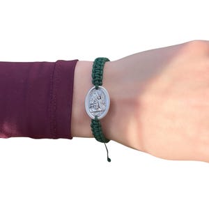 May include: A green braided bracelet with a silver oval charm featuring a religious image. The charm has a raised design of an angel with wings.