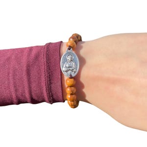 May include: A brown wooden bead bracelet with a silver oval charm featuring a religious figure. The charm has the words "St. Jude" inscribed on it.