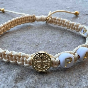 Puede incluir: Un brazalete trenzado blanco con una medalla de San Cristóbal de oro y cuentas blancas con la palabra "PAX" escrita en letras doradas.