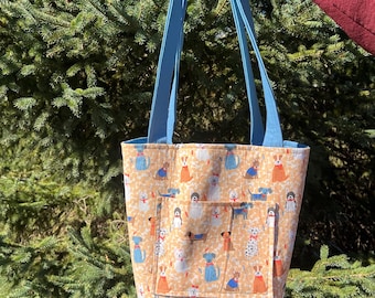 Può includere: Una borsa tote trapuntata con sfondo marrone chiaro e un motivo ripetuto di cani in vari abiti. La borsa ha una tasca frontale e cinghie azzurre. La borsa è tenuta contro uno sfondo di fogliame verde.