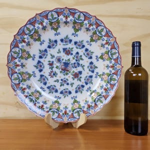 May include: A decorative white ceramic plate with a floral design in blue, red, and green. The plate has a scalloped edge and is displayed on a wooden stand. A brown wine bottle is next to the plate.