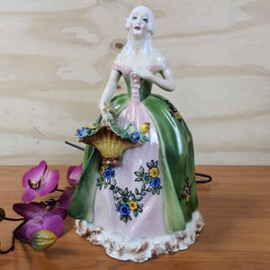 Może przedstawiać: Porcelanowa figurka kobiety w długiej zielono-różowej sukni, trzymającej kosz kwiatów. Suknia jest ozdobiona kwiatowymi wzorami. Figurka ma białą perukę i stoi na ozdobnej podstawie.