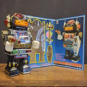 Könnte beinhalten: Ein schwarzer und silberner Spielzeugroboter mit einer durchsichtigen Plastikbrust und einem roten Licht oben. Der Roboter hält eine silberne Pistole in seiner rechten Hand. Der Roboter steht auf einer Holzoberfläche. Die Schachtel für den Roboter ist geöffnet und zeigt den Roboter in einer anderen Pose. Die Schachtel ist blau und weiß und hat den Schriftzug "STAR ROTO ROBOT" aufgedruckt.
