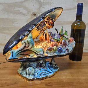 Könnte beinhalten: Eine Keramiklampe in Muschelform mit dunkelblauem Äußeren und Goldrand, die ein beleuchtetes Inneres mit geformten Korallen, Algen und bunten Fischfiguren zeigt. Eine dunkle Glasweinflasche steht neben der Lampe auf einer Holzoberfläche.