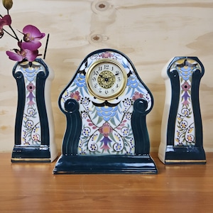 May include: A decorative ceramic clock and matching vases. The clock has a gold-tone face with black numerals and hands. The clock and vases feature a floral pattern in blue, pink, and purple on a white background, with dark green trim.