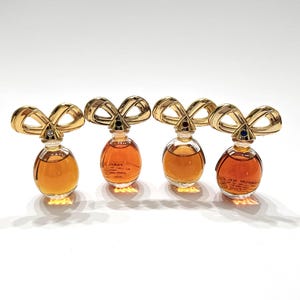 May include: Four miniature perfume bottles with clear glass bodies containing amber liquid. Each bottle features a gold-toned metal cap shaped like a stylized bow, accented with a small, colored gemstone at the center of the bow knot.