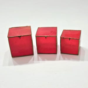 May include: Three red square boxes with brass-colored metal trim and hinged lids. The boxes are arranged in a row, decreasing in size from left to right. The boxes are on a white surface.