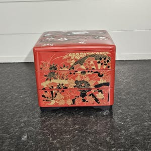 May include: A red, three-tiered Japanese bento box with black and gold floral and fan designs. The box has a glossy finish and sits on a black base. The top of the box also features white floral accents.