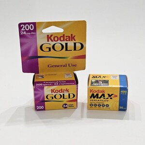 May include: Three boxes of Kodak film are stacked. The top box is a yellow and purple Kodak Gold film package with the text "200 24 exp. film." Below it is a box of Kodak Gold 200 speed film. To the right is a blue and yellow Kodak Max film box.