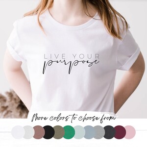 Puede incluir: Camiseta blanca con el texto "LIVE YOUR purpose" en letra cursiva negra. La camiseta se muestra en una modelo. Más colores para elegir se muestran debajo de la modelo.