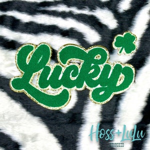 May include: Green felt "Lucky" lettering with gold glitter trim and a gold glitter shamrock on a black and white zebra print background. The text "Hoss + Lulu Stitched" is also visible.