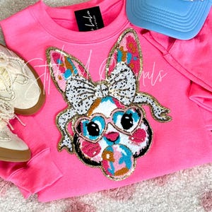 May include: Pink sweatshirt with a large sequined bunny applique. The bunny is wearing heart-shaped sunglasses and a bow. The sweatshirt has a black label with the text "Little Lulu" on it.