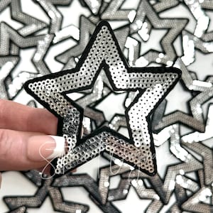 May include: A silver sequin star-shaped patch with a black outline. The patch is made of sequins and has a shiny finish.