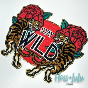 May include: An embroidered patch featuring two tigers and roses in red, gold and black. The text "Stay Wild" is in the centre of the patch.