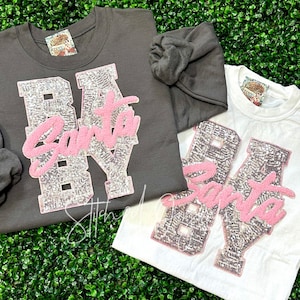 May include: Two gray and white t-shirts with the text "By Santa" in pink glitter letters. The letters are outlined in black and have a sparkly silver finish.
