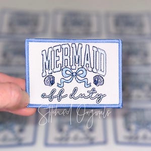 May include: Embroidered patch with a white background and a light blue border. The patch features the words "MERMAID off duty" in blue lettering, along with a bow and two seashell designs. The patch is rectangular.