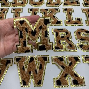 May include: A set of brown and black felt letters with gold trim. The letters are in a leopard print pattern and are arranged in alphabetical order. The letters are M, R, S, W, and X.