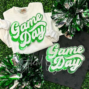 May include: Two sweatshirts with the words "Game Day" in green and white sequins. The sweatshirts are on a green background with green and silver pom-poms.