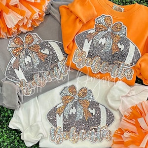 May include: Three white shirts with a black and white football design with rhinestones. The football has a brown bow on top. The shirts are layered on top of each other.