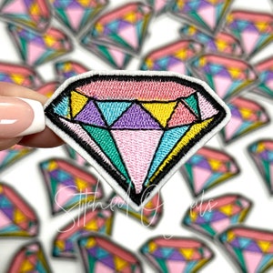 May include: Embroidered diamond patch with a colorful design. The patch features a pink, purple, yellow, and teal gemstone design with a black outline and white border. The patch is held up in front of a blurred background of similar patches.
