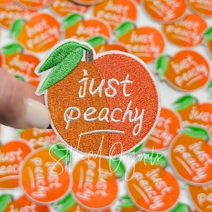 May include: A close-up of an embroidered iron-on patch with a peach design and the text "just peachy".