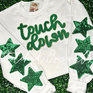 May include: White sweatshirt with the words "touch down" in green and gold glitter lettering. The sleeves have green sequin stars.