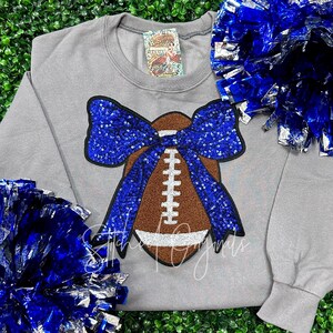 May include: Gray sweatshirt with a brown football and a blue sequined bow. The football has white stitching and the bow has a blue sequined center.