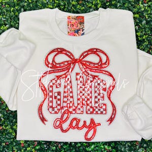 May include: A white sweatshirt with long sleeves. The front features the words "GAME DAY" in red sequined letters, with a large red bow above. The letters have a checkered pattern. The sweatshirt is on a green background.