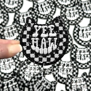 May include: A black and white checkered iron-on patch with the text "Yee Haw" in white embroidery. The patch is shaped like a smiling cat face.