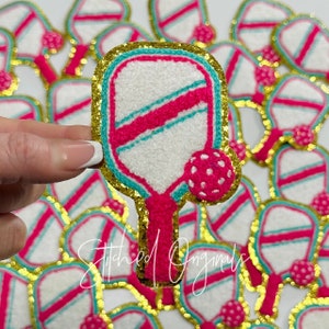 May include: A collection of colorful pickleball paddle patches. Each patch features a white paddle with a pink and teal design, a pink ball, and a gold glitter border. The text "Stitched Originals" is written in cursive on the bottom.