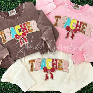 May include: Three sweatshirts in brown, pink, and cream, each with the word "TEACHER" in colorful sequined letters and a red bow. The sweatshirts have ribbed cuffs and collars. The text "Stitched Originals" is also visible.
