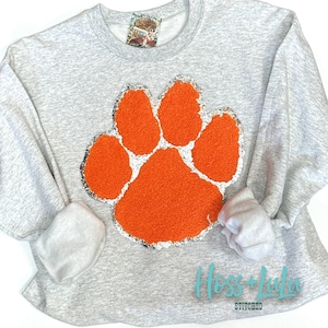 May include: Gray sweatshirt with a large orange paw print design. The paw print is made of a fuzzy material and has a silver glitter outline. The sweatshirt has the text "Hoss + Lula Stitched" in teal blue lettering on the bottom.