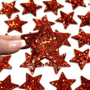 May include: A close-up of a hand holding a large, orange, sparkly star-shaped patch. The patch is made of sequins and is surrounded by other smaller, orange, sparkly star-shaped patches.