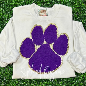 May include: White sweatshirt with a purple and gold paw print design. The paw print is outlined in gold glitter and the inside is a fuzzy purple material.
