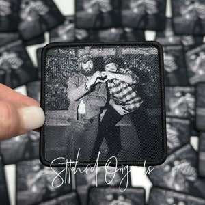 May include: Black and white square patch featuring a photograph of two people making a heart shape with their hands. The patch has a black border and the words "Stitched Ong's" in cursive script. Patches are scattered in the background.