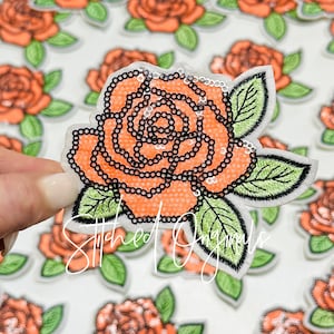 May include: A peach-colored rose with black and white sequins and green leaves. The rose is embroidered on a white fabric.