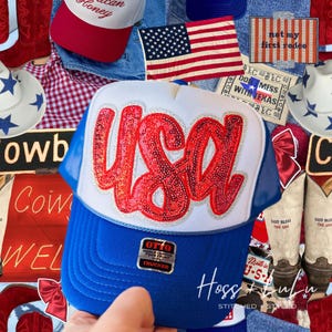 May include: A blue and white trucker hat with the word "USA" in red sequins. The hat has a blue brim and mesh backing. The image also includes a US flag, cowboy boots, and other patriotic themed items.
