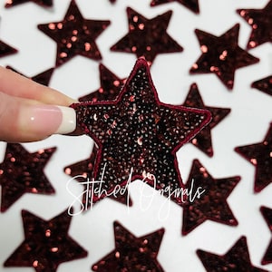 May include: A close-up of a burgundy sequined star-shaped patch. The patch is surrounded by other similar patches, all on a white background.