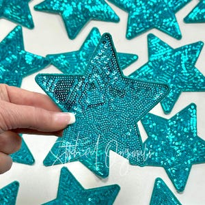 May include: Turquoise sequin star patches. The patches are embroidered with small, shiny sequins. The text "Stitched Originals" is visible on one of the stars. These patches can be used for various craft projects.