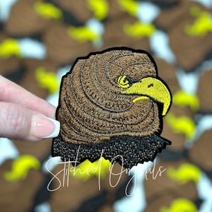 May include: Brown and yellow embroidered eagle head patch with black stitching. The patch has a jagged edge and is held by a hand.