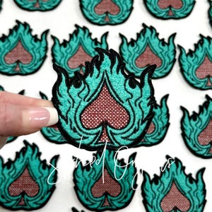 May include: A set of teal and red embroidered iron-on patches featuring a stylized playing card spade in flames.