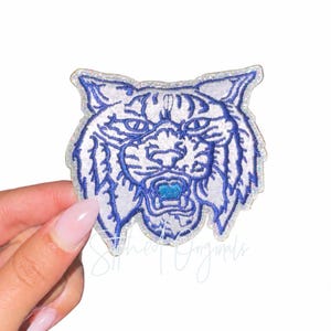 May include: An embroidered patch featuring the head of a tiger with white and silver glitter thread details and bold blue stitching for the facial features, whiskers, and ears. The tiger has an open mouth with a bright blue center accent.