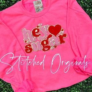 May include: Pink sweatshirt with a red and pink glitter design that says "Hey Sugar" with a red heart in the middle. The sweatshirt has the words "Stitched Originals" written in white on the bottom.