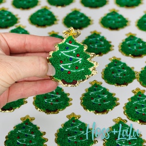 May include: A close-up of a hand holding a green and gold glitter Christmas tree patch. The patch is embroidered with white and red details. The patch is surrounded by other Christmas tree patches.