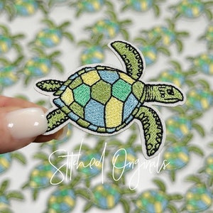 May include: A green, blue, and yellow embroidered sea turtle patch. The turtle is outlined in black thread and has a detailed shell pattern.