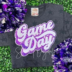 May include: A dark gray t-shirt with a white and purple sequined "Game Day" design. The shirt has a distressed look and is decorated with small white stars.