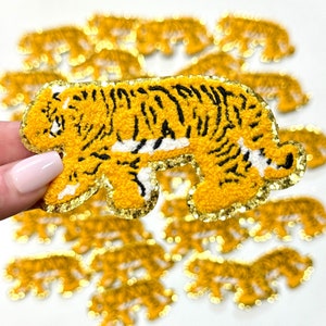 May include: A close-up of a yellow and black tiger applique with glitter trim. The applique is made of a fuzzy fabric and is perfect for adding a touch of whimsy to any project.