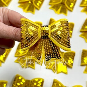 May include: A close-up of a gold sequined bow with a stitched edge. The bow is made of small, round sequins that are sewn together to create a shimmering effect.