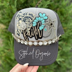 Op de afbeelding: Grijze truckercap met een turquoise en zilveren versiering, een patch van een cowboy die een bokkend bronco berijdt, en het woord "faith" in zwarte en gouden letters. De pet heeft ook een rand van strass en de woorden "Stitched Originals" op de klep.